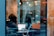 A person is seated at a table inside a cafe, working on a laptop with documents open on the screen. Nearby, a smartphone is propped up, perhaps for video calls. The window reflects a person walking outside and the urban street scene with parked cars.