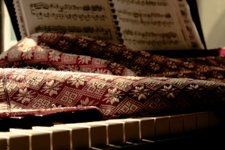 A sheet of music rests on a stand above a piano keyboard. A textured fabric with intricate patterns covers parts of the piano, with sunlight casting warm tones and soft shadows over the scene.