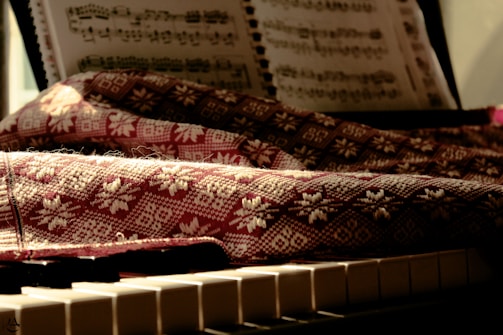 A sheet of music rests on a stand above a piano keyboard. A textured fabric with intricate patterns covers parts of the piano, with sunlight casting warm tones and soft shadows over the scene.
