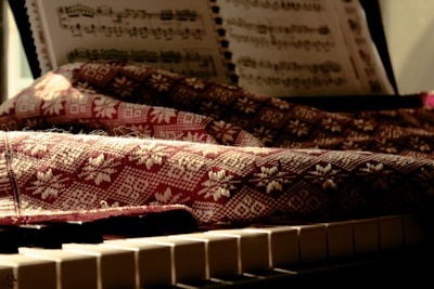 A sheet of music rests on a stand above a piano keyboard. A textured fabric with intricate patterns covers parts of the piano, with sunlight casting warm tones and soft shadows over the scene.
