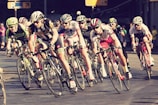 A group of cyclists in racing attire are intensely competing in a road race. They are in close proximity to each other, leaning into a turn on a paved street. The setting appears to be urban with some spectators in the background. Bicycle wheels and handlebars are prominent, showcasing a sense of speed and competition.