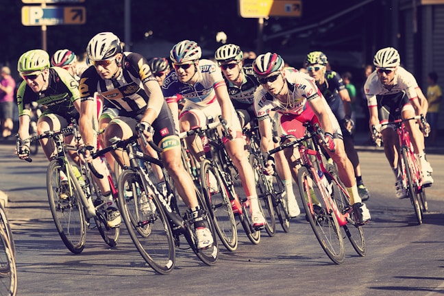 A group of cyclists in racing attire are intensely competing in a road race. They are in close proximity to each other, leaning into a turn on a paved street. The setting appears to be urban with some spectators in the background. Bicycle wheels and handlebars are prominent, showcasing a sense of speed and competition.