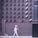 A person wearing a helmet with a smiley face is walking along a wall covered with numerous surveillance cameras pointed in various directions. The wall is made of bricks with a minimalist design, and the cameras are arranged in neat rows.