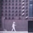 A person wearing a helmet with a smiley face is walking along a wall covered with numerous surveillance cameras pointed in various directions. The wall is made of bricks with a minimalist design, and the cameras are arranged in neat rows.