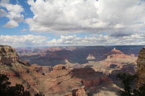 A vast and majestic canyon landscape with layered rock formations in various shades of red and brown. The sky overhead is partially covered with white, fluffy clouds, allowing patches of sunlight to illuminate parts of the canyon floor, creating contrasting light and shadow effects.