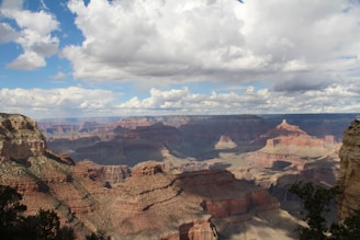 A vast and majestic canyon landscape with layered rock formations in various shades of red and brown. The sky overhead is partially covered with white, fluffy clouds, allowing patches of sunlight to illuminate parts of the canyon floor, creating contrasting light and shadow effects.