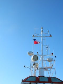 A ship's mast is prominently featured against a clear blue sky. The mast is equipped with various radar and communication devices. A red, white, and blue flag is waving at the top.