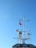 A ship's mast is prominently featured against a clear blue sky. The mast is equipped with various radar and communication devices. A red, white, and blue flag is waving at the top.