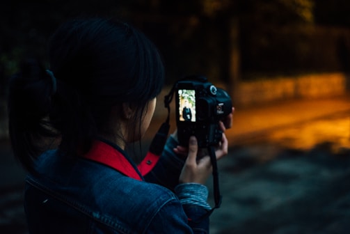 A person with dark hair pulled back is holding a DSLR camera, capturing an image on a dimly lit street. The camera's screen displays a smaller version of the photo being taken, reflecting a figure standing at a distance.