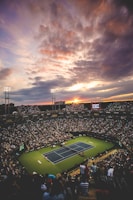 A large crowd fills a tennis stadium at sunset, with a match taking place on the court. The sky is dramatic, filled with clouds illuminated by the setting sun, creating a picturesque backdrop. Stadium lights are turned on, enhancing the vibrant atmosphere.