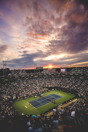 A large crowd fills a tennis stadium at sunset, with a match taking place on the court. The sky is dramatic, filled with clouds illuminated by the setting sun, creating a picturesque backdrop. Stadium lights are turned on, enhancing the vibrant atmosphere.