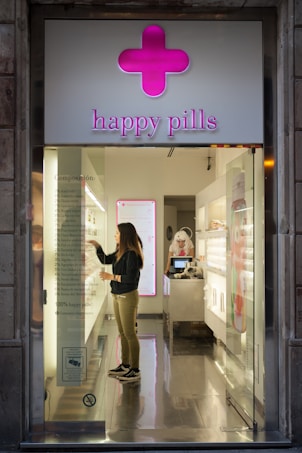 A store with a modern design and bright pink signage labeled 'happy pills' features a woman standing inside, examining the contents of a transparent display. The interior is sleek and glossy with a bright, organized layout. Another person, dressed in white, is visible behind the counter, which displays an array of colorful items.