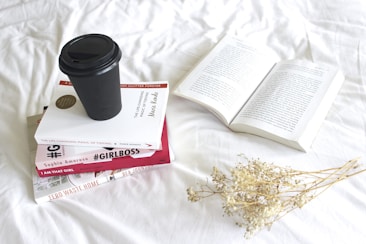 A stack of five books with a black disposable coffee cup on top is placed on a white surface. Next to the stack is an open book, and to the side lies a small bunch of dried flowers.