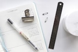 An open planner with a dotted design displays future logs for the months of July and August. A black pen, a metal bulldog clip, a ruler, and two safety pins are placed atop and beside the planner. A small container with a marble-like texture is partially visible.