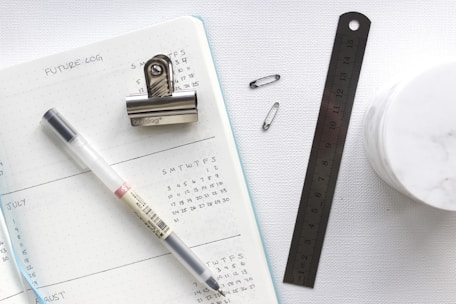 An open planner with a dotted design displays future logs for the months of July and August. A black pen, a metal bulldog clip, a ruler, and two safety pins are placed atop and beside the planner. A small container with a marble-like texture is partially visible.