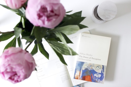 A soft-focused composition featuring a book titled 'The Doors of Perception and Heaven and Hell' by Aldous Huxley placed on a white surface. Surrounding the book are three pink peonies with green leaves, and another open book or planner with visible text. A marble or ceramic round object is partially visible near the top right of the image.