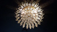 A luxurious and ornate crystal chandelier with intricate glass and metalwork. The chandelier is illuminated, casting a warm glow and creating shimmering reflections on the surrounding surfaces. It has an elaborate, almost sunburst design with numerous faceted crystal pieces radiating outward.