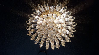 A luxurious and ornate crystal chandelier with intricate glass and metalwork. The chandelier is illuminated, casting a warm glow and creating shimmering reflections on the surrounding surfaces. It has an elaborate, almost sunburst design with numerous faceted crystal pieces radiating outward.