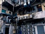 An industrial setting with numerous air conditioning units and large metallic pipes attached to the exterior of a weathered, multi-story building. The walls show signs of wear, with peeling paint and visible stains. The area appears cluttered with wiring and equipment, suggesting a functional but neglected environment.