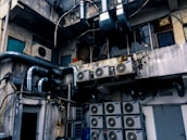 An industrial setting with numerous air conditioning units and large metallic pipes attached to the exterior of a weathered, multi-story building. The walls show signs of wear, with peeling paint and visible stains. The area appears cluttered with wiring and equipment, suggesting a functional but neglected environment.