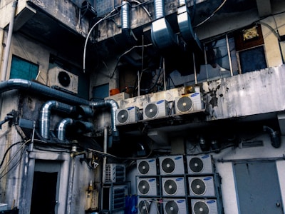 An industrial setting with numerous air conditioning units and large metallic pipes attached to the exterior of a weathered, multi-story building. The walls show signs of wear, with peeling paint and visible stains. The area appears cluttered with wiring and equipment, suggesting a functional but neglected environment.