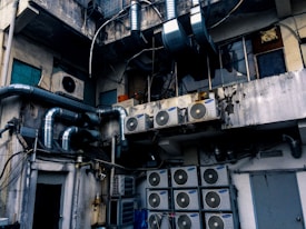 An industrial setting with numerous air conditioning units and large metallic pipes attached to the exterior of a weathered, multi-story building. The walls show signs of wear, with peeling paint and visible stains. The area appears cluttered with wiring and equipment, suggesting a functional but neglected environment.