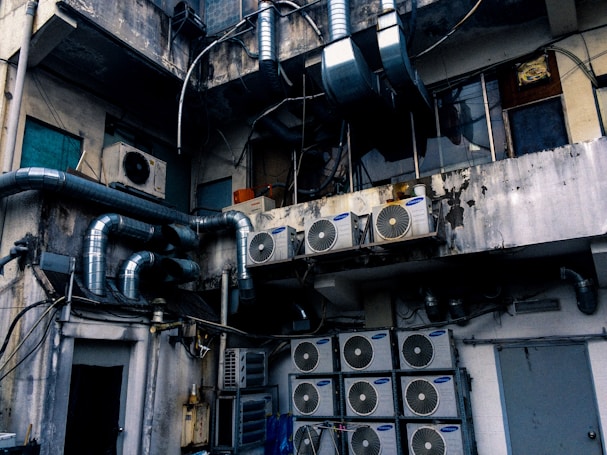 An industrial setting with numerous air conditioning units and large metallic pipes attached to the exterior of a weathered, multi-story building. The walls show signs of wear, with peeling paint and visible stains. The area appears cluttered with wiring and equipment, suggesting a functional but neglected environment.