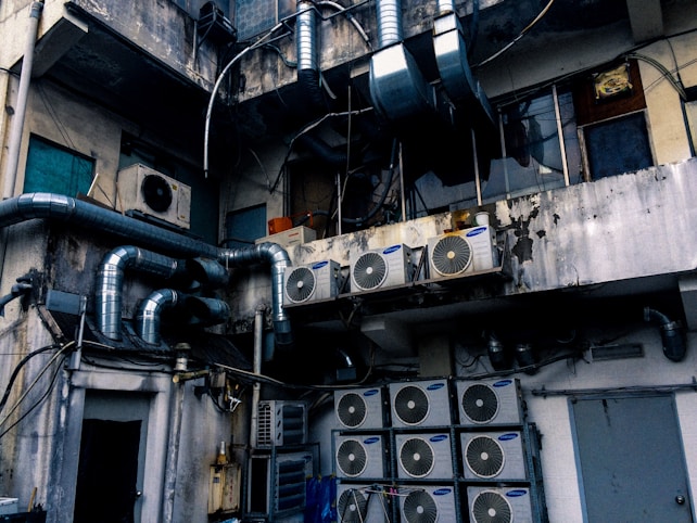 An industrial setting with numerous air conditioning units and large metallic pipes attached to the exterior of a weathered, multi-story building. The walls show signs of wear, with peeling paint and visible stains. The area appears cluttered with wiring and equipment, suggesting a functional but neglected environment.