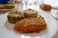 A close-up view of a white plate containing three differently textured and colored food items: one appears green and leafy, another is golden brown and crispy, and the third is a mixture of white and darker grains, likely quinoa. In the background, there is a glass of water and a loaf of bread on the table.