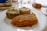 A close-up view of a white plate containing three differently textured and colored food items: one appears green and leafy, another is golden brown and crispy, and the third is a mixture of white and darker grains, likely quinoa. In the background, there is a glass of water and a loaf of bread on the table.