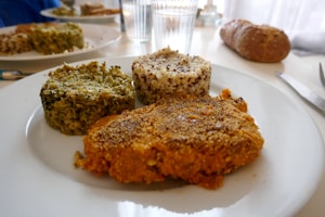 A close-up view of a white plate containing three differently textured and colored food items: one appears green and leafy, another is golden brown and crispy, and the third is a mixture of white and darker grains, likely quinoa. In the background, there is a glass of water and a loaf of bread on the table.