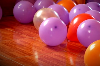 A group of colorful balloons spread out on a shiny wooden floor, featuring mainly purple, orange, and red hues. Light reflects off the smooth surfaces of the balloons, creating a festive and vibrant atmosphere.