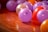 A group of colorful balloons spread out on a shiny wooden floor, featuring mainly purple, orange, and red hues. Light reflects off the smooth surfaces of the balloons, creating a festive and vibrant atmosphere.