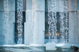 Water cascades in streams over large stone slabs, creating a textured pattern as it flows into a shallow basin below. The scene has an architectural feel with the structured placement of rough stone surfaces and clear displays of water movement.