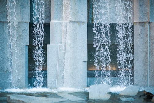 Water cascades in streams over large stone slabs, creating a textured pattern as it flows into a shallow basin below. The scene has an architectural feel with the structured placement of rough stone surfaces and clear displays of water movement.