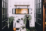 An outdoor space with a focus on gardening, featuring green ivy plants climbing up on two black pillars. A weathered watering can hangs prominently as part of the decor. Above, a wooden shelf holds wire baskets and a decorative bird. The scene includes lanterns suspended from the ceiling and a centered mirror reflecting an indoor space.
