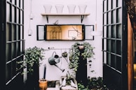 An outdoor space with a focus on gardening, featuring green ivy plants climbing up on two black pillars. A weathered watering can hangs prominently as part of the decor. Above, a wooden shelf holds wire baskets and a decorative bird. The scene includes lanterns suspended from the ceiling and a centered mirror reflecting an indoor space.