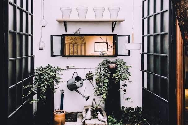 An outdoor space with a focus on gardening, featuring green ivy plants climbing up on two black pillars. A weathered watering can hangs prominently as part of the decor. Above, a wooden shelf holds wire baskets and a decorative bird. The scene includes lanterns suspended from the ceiling and a centered mirror reflecting an indoor space.