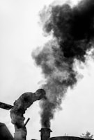 A silhouetted figure leans forward near a thick plume of black smoke rising from a chimney or exhaust. The scene is in black and white, emphasizing the contrast between the dark smoke and the lighter background.