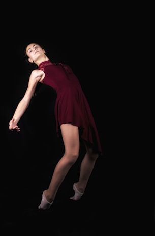 A dancer in a burgundy dress is captured in a graceful pose against a dark background. She is leaning back with an elegant arch, displaying balance and poise. Her arms are extended, contributing to the fluidity and motion of the dance.