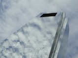 A modern skyscraper with a reflective glass facade captures the sky and clouds, creating an illusion of transparency and openness. The structure rises at an angle, emphasizing its height and architectural design.