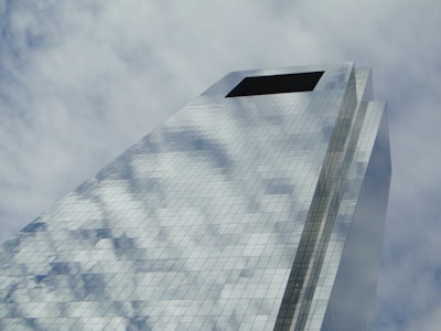A modern skyscraper with a reflective glass facade captures the sky and clouds, creating an illusion of transparency and openness. The structure rises at an angle, emphasizing its height and architectural design.