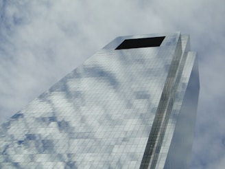 A modern skyscraper with a reflective glass facade captures the sky and clouds, creating an illusion of transparency and openness. The structure rises at an angle, emphasizing its height and architectural design.