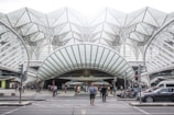 A modern transportation hub with an intricate steel and glass architectural design. People are walking across the street, surrounded by several vehicles including cars and a motorcycle. The structure features a series of overlapping geometric patterns, creating an open and airy atmosphere.