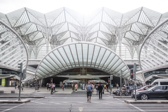 A modern transportation hub with an intricate steel and glass architectural design. People are walking across the street, surrounded by several vehicles including cars and a motorcycle. The structure features a series of overlapping geometric patterns, creating an open and airy atmosphere.