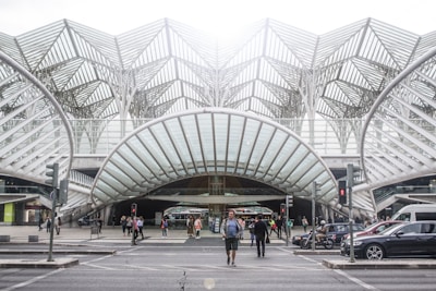 A modern transportation hub with an intricate steel and glass architectural design. People are walking across the street, surrounded by several vehicles including cars and a motorcycle. The structure features a series of overlapping geometric patterns, creating an open and airy atmosphere.