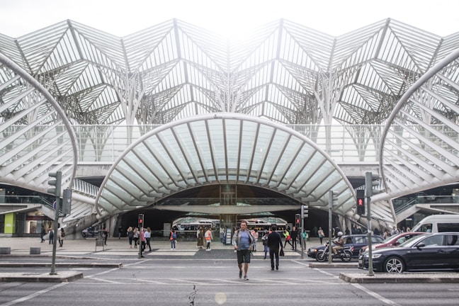 A modern transportation hub with an intricate steel and glass architectural design. People are walking across the street, surrounded by several vehicles including cars and a motorcycle. The structure features a series of overlapping geometric patterns, creating an open and airy atmosphere.