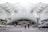 A modern transportation hub with an intricate steel and glass architectural design. People are walking across the street, surrounded by several vehicles including cars and a motorcycle. The structure features a series of overlapping geometric patterns, creating an open and airy atmosphere.
