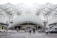 A modern transportation hub with an intricate steel and glass architectural design. People are walking across the street, surrounded by several vehicles including cars and a motorcycle. The structure features a series of overlapping geometric patterns, creating an open and airy atmosphere.