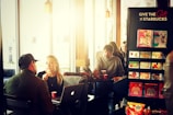 Several people are seated in a coffee shop with large windows allowing in natural light. A man and woman are engaged in conversation at a table, where a laptop is open in front of the woman. The man is wearing a hat and a jacket, while the woman has long blonde hair and is holding a small red cup. A display stand offers Starbucks gift cards. In the background, other patrons are sitting at tables enjoying their beverages.